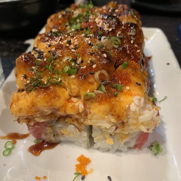 Close-up of a sushi roll with baked topping, sesame seeds, and green onions on a white plate.